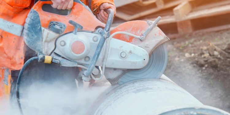 Construction worker in orange safety gear uses a handheld power saw to cut a large metal pipe, creating clouds of dust.